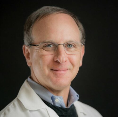 A person with dark brown and gray hair standing in front of a black background wearing a lab coat and black sweater.