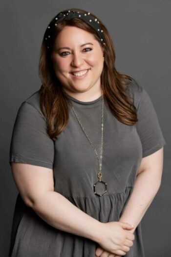 A person with long brown hair wearing a dark gray dress and gold necklace stands smiling in front of a gray background.
